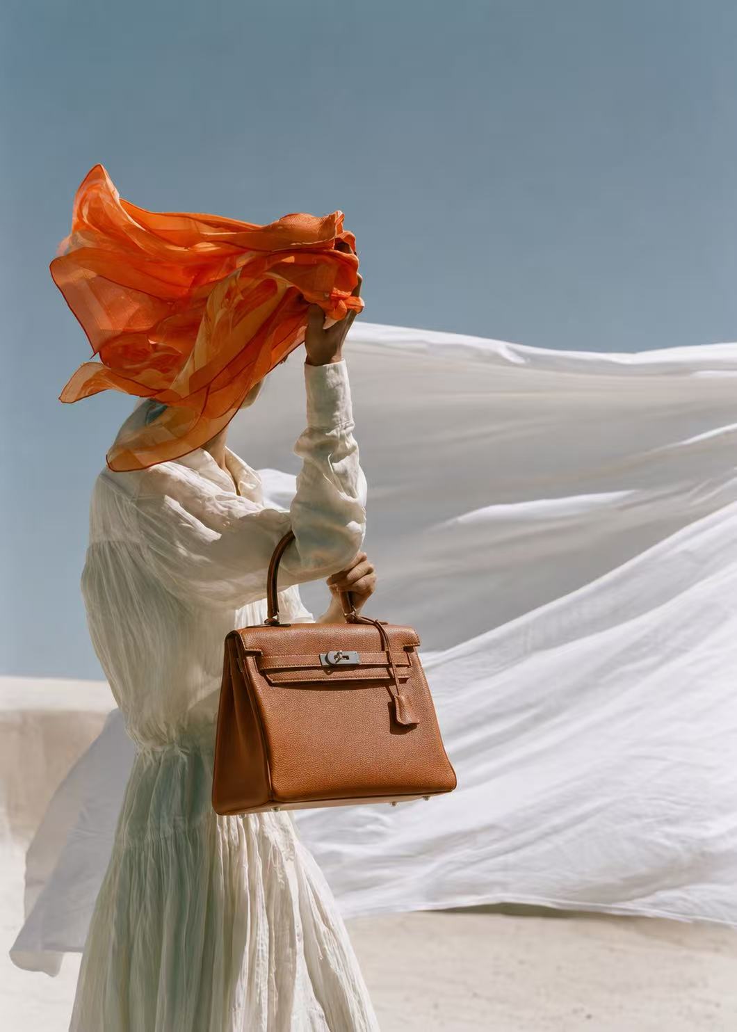 A woman in white holding a tan Hermès Kelly bag against flowing fabric and blue sky